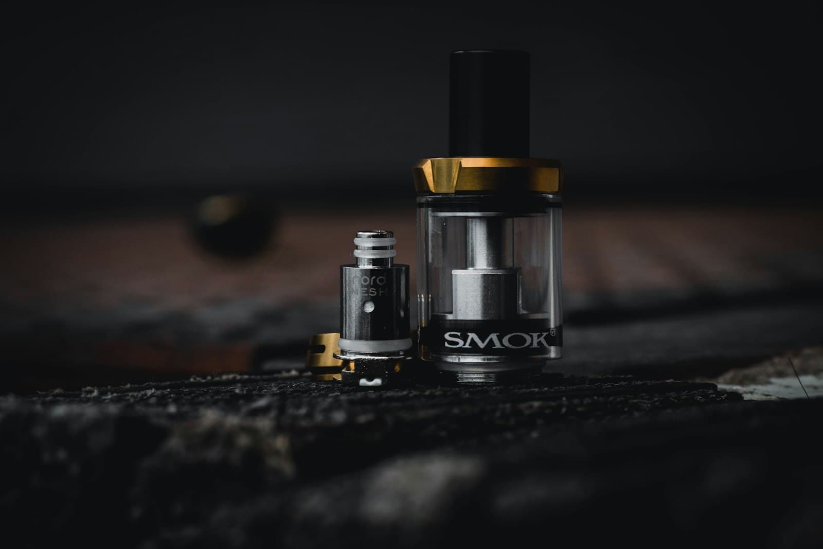 A clear sub-ohm vape tank standing next to its mesh replacement coil on a dark wooden surface